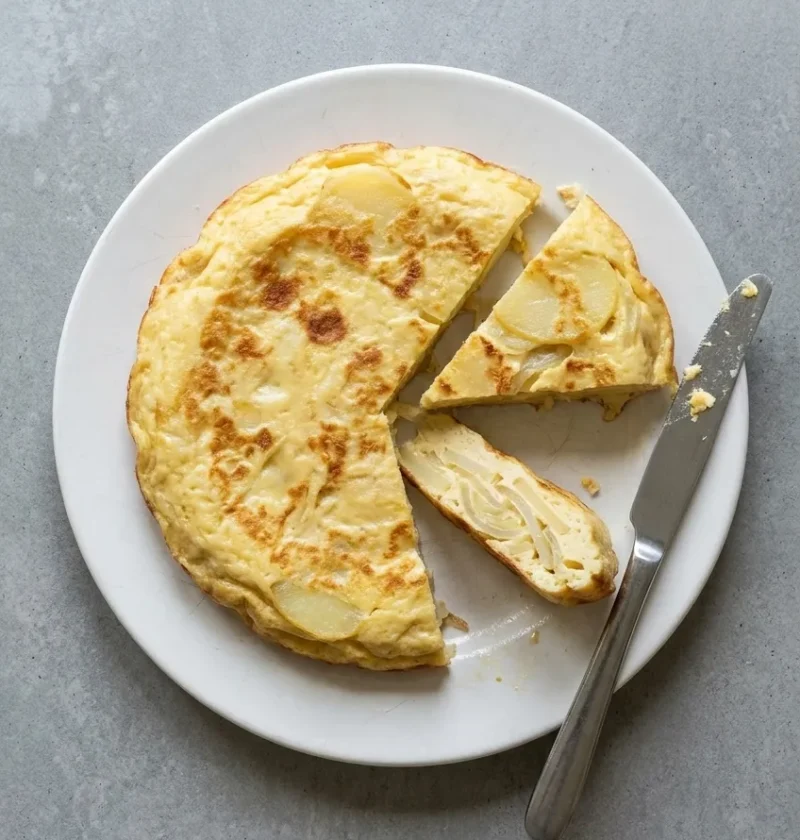 A whole Spanish tortilla on a white plate with two wedges cut out, showing thin slices of potato and onion set in pale golden egg