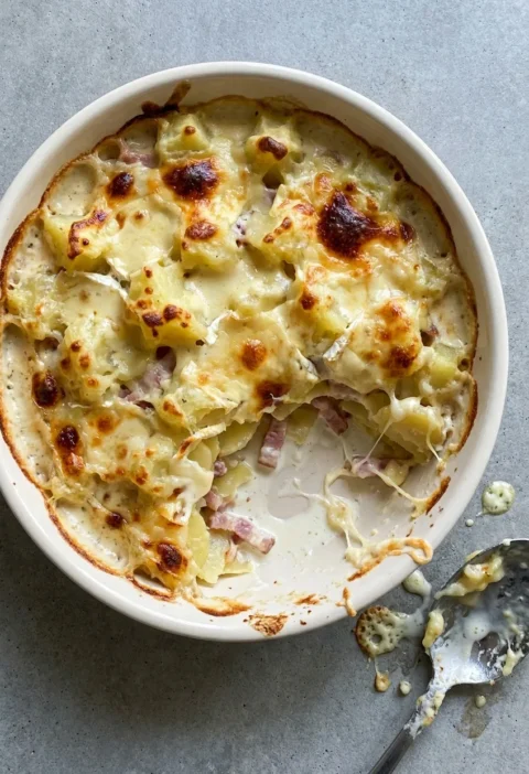 Hairy Bikers tartiflette in a white dish, melted reblochon over potatoes and bacon with a scoop taken out