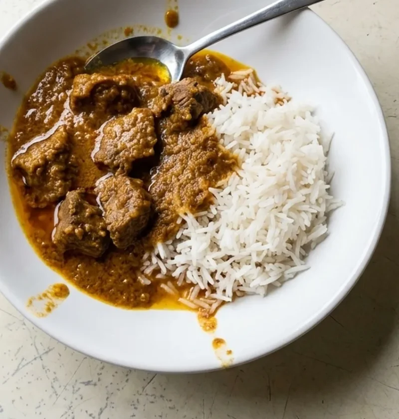 A white bowl of beef curry with tender chunks of braising steak in a spiced tomato and yoghurt sauce, served with basmati rice and a spoon