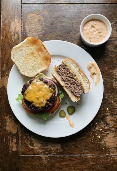 Homemade beef burger with melted Cheddar and a halved burger on a white plate, with a ramekin of burger sauce, on a worn oak table