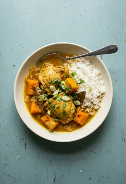 A bowl of Caribbean chicken curry with skinless chicken thighs, pumpkin chunks, thyme and a bay leaf in a golden sauce, with steamed rice