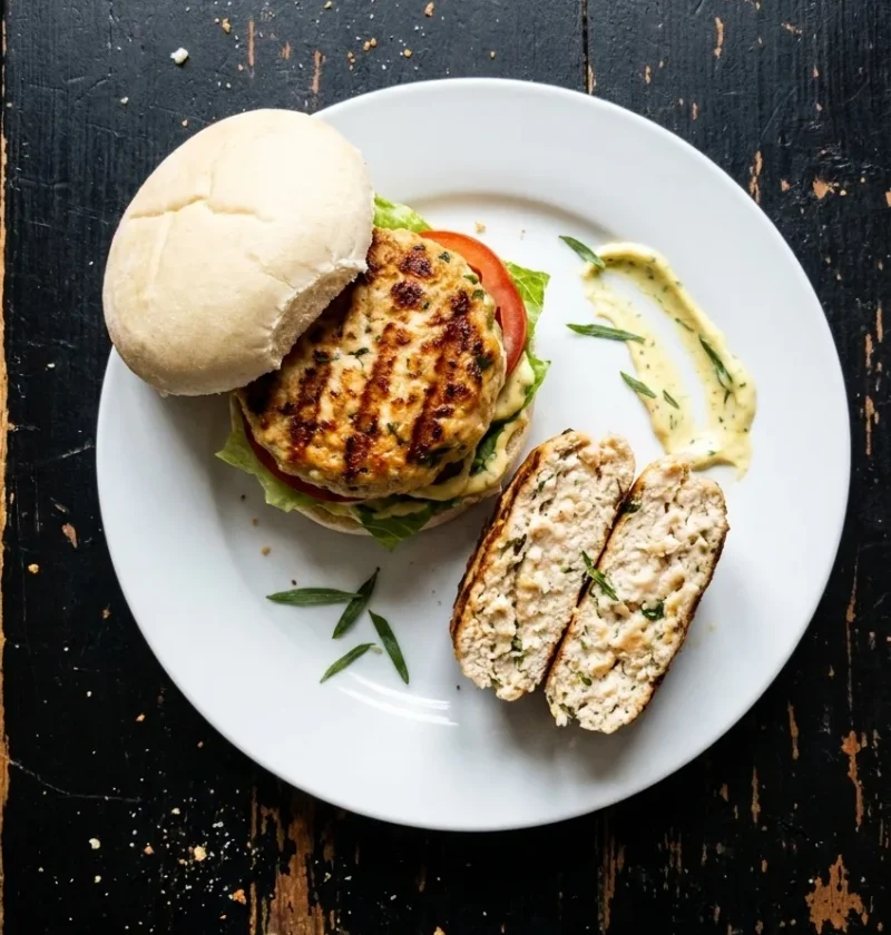Griddled chicken burger with lettuce and tomato in a soft bun, beside a halved patty and a smear of tarragon mayo, on a worn black table