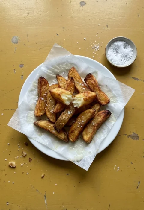 Triple-cooked chips piled on kitchen paper on a white plate, one snapped open, with a dish of flaked sea salt