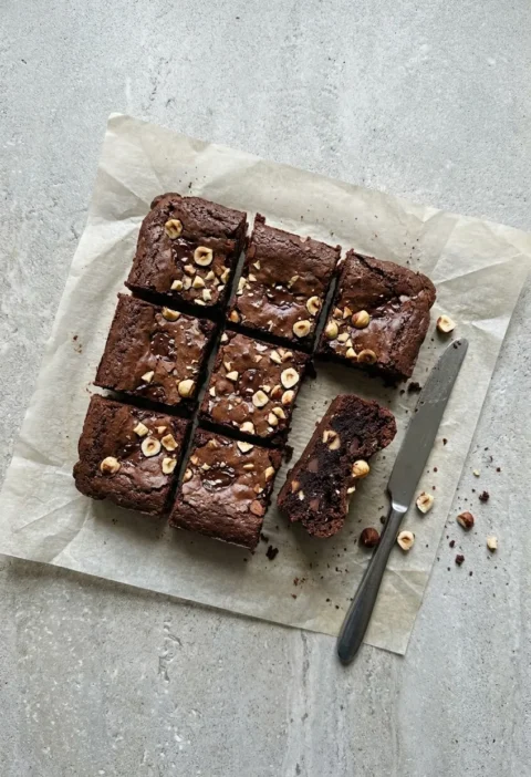 Squares of dark chocolate hazelnut brownies on baking paper, topped with chopped hazelnuts, one on its side showing the fudgy inside