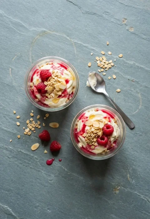 Two glass tumblers of cranachan from above, whipped cream rippled with raspberry purée and topped with raspberries, toasted oats and flaked almonds
