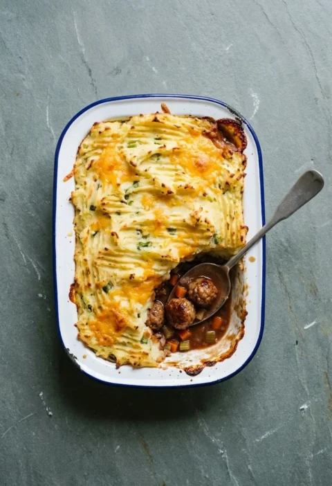 Cumberland sausage pie in an enamel dish with golden forked cheesy mash, one corner scooped out to show sausage balls in red wine gravy