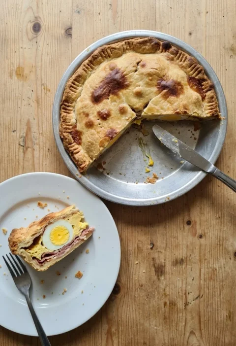 Homemade egg and bacon pie in a metal pie tin on a pine table, with a slice on a plate showing a whole set egg and bacon inside