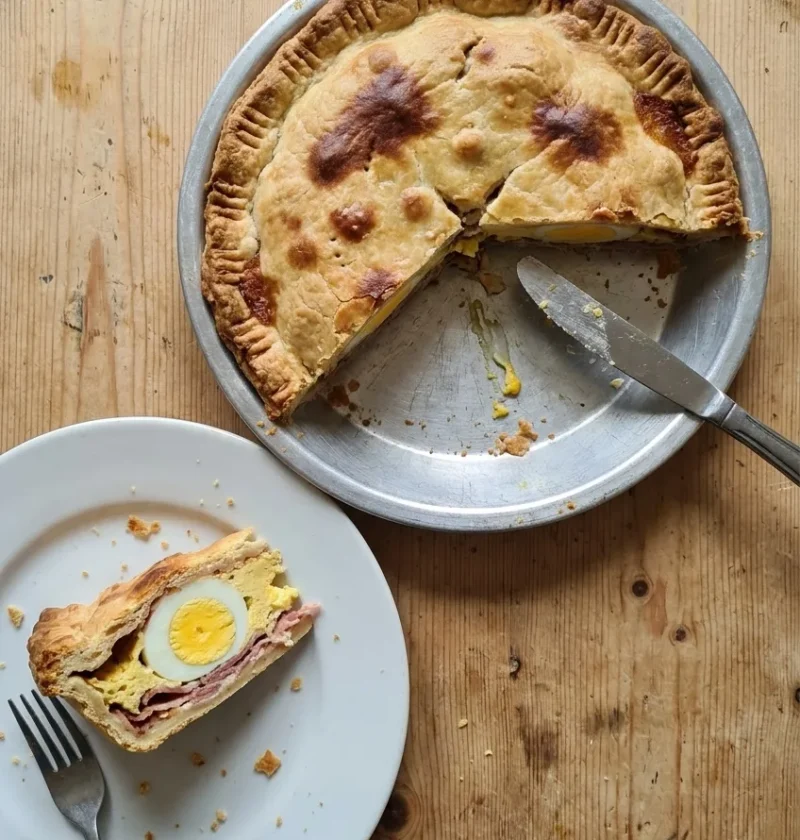 Homemade egg and bacon pie in a metal pie tin on a pine table, with a slice on a plate showing a whole set egg and bacon inside