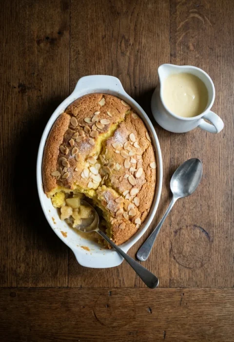 A white oval dish of Eve’s pudding with an almond-topped sponge, one portion scooped out to show soft apple, beside a jug of vanilla custard