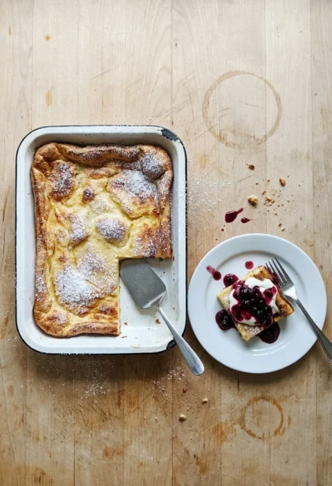 Finnish oven pancake in an enamel baking dish dusted with icing sugar, one square served with berry compote and crème fraîche
