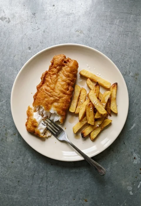 A plate of beer-battered haddock broken open to show white flaky fish, with thick-cut chips sprinkled with salt