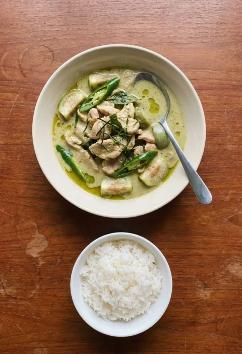 A bowl of Thai green chicken curry with apple aubergines, green chillies, lime leaf strips and Thai basil, with a small bowl of jasmine rice
