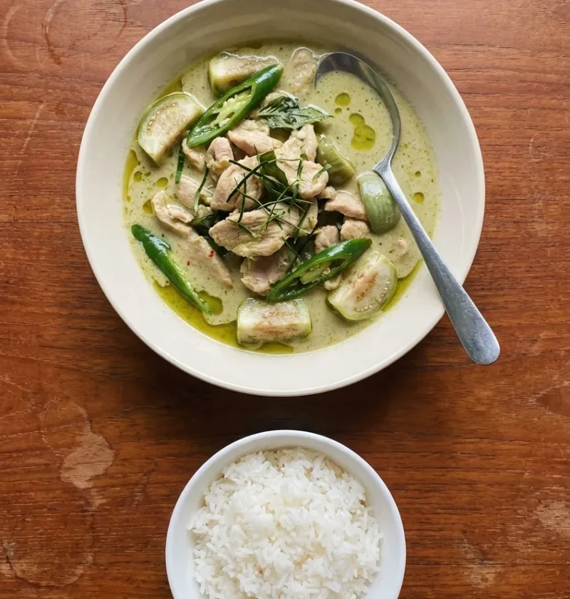 A bowl of Thai green chicken curry with apple aubergines, green chillies, lime leaf strips and Thai basil, with a small bowl of jasmine rice
