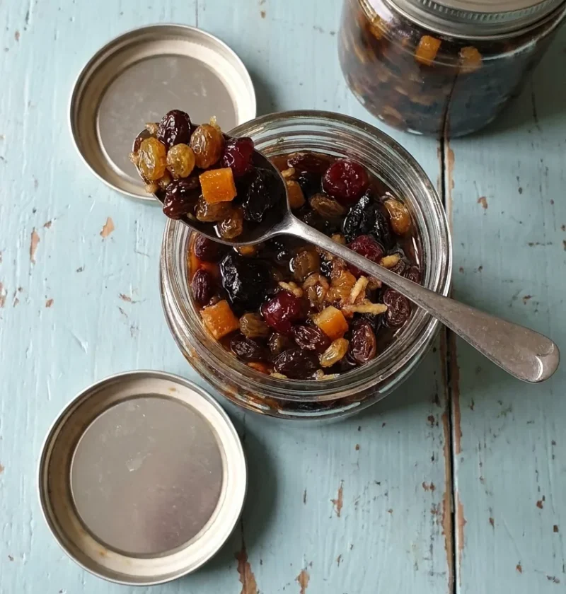 Hairy Bikers mincemeat in an open glass jar with a spoonful of raisins, sultanas, sour cherries and candied orange peel, a second jar behind