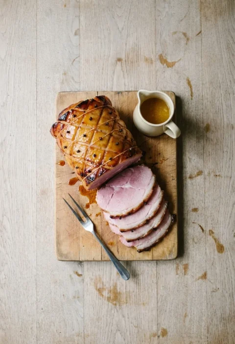 A honey and mustard glazed gammon joint studded with cloves on a carving board, with thick pink slices and a jug of warm juices