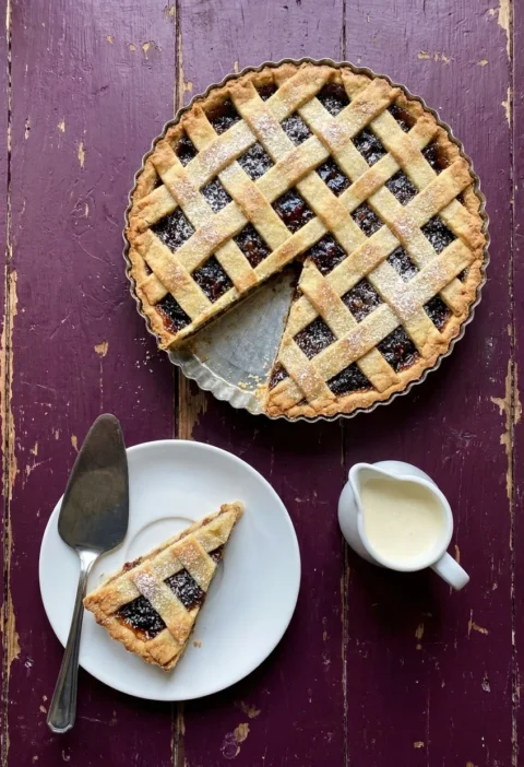 Hairy Bikers mincemeat tart with a golden almond pastry lattice in a fluted tin, one slice served on a white plate with a jug of cream