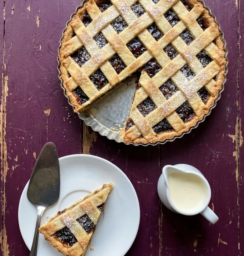 Hairy Bikers mincemeat tart with a golden almond pastry lattice in a fluted tin, one slice served on a white plate with a jug of cream