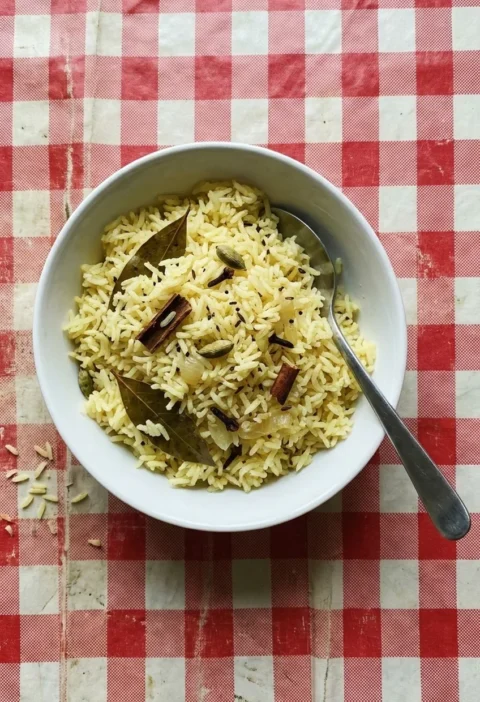 A white bowl of pale yellow pilau rice with whole cardamom pods, cinnamon, cloves and bay leaves, with a serving spoon, on a red checked oilcloth