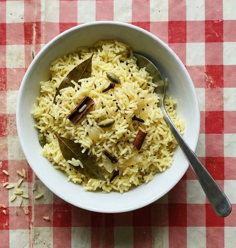 A white bowl of pale yellow pilau rice with whole cardamom pods, cinnamon, cloves and bay leaves, with a serving spoon, on a red checked oilcloth