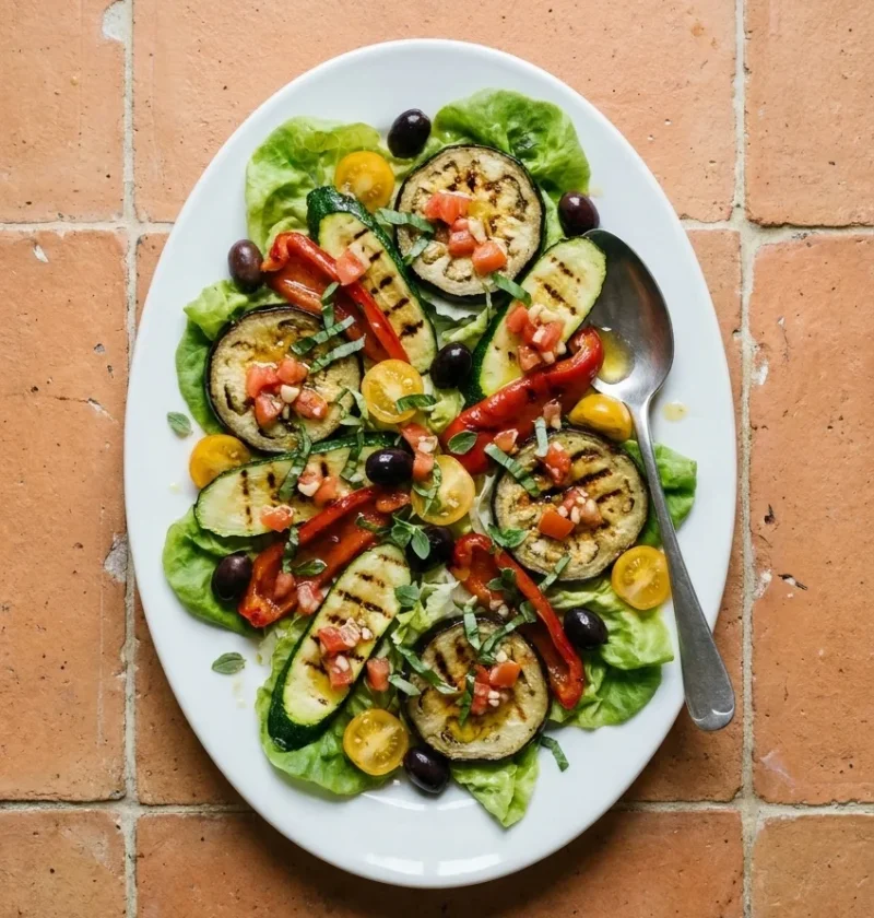 A white platter of griddled aubergine and courgette slices with roast red pepper, yellow cherry tomatoes, black olives and a diced tomato dressing