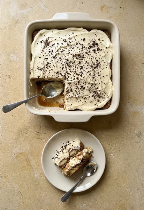 A tiramisu in a cream baking dish topped with fine dark chocolate shards, one corner spooned out, with a portion on a small plate showing coffee-soaked sponge fingers