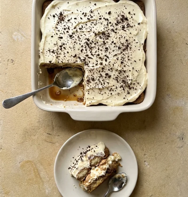 A tiramisu in a cream baking dish topped with fine dark chocolate shards, one corner spooned out, with a portion on a small plate showing coffee-soaked sponge fingers