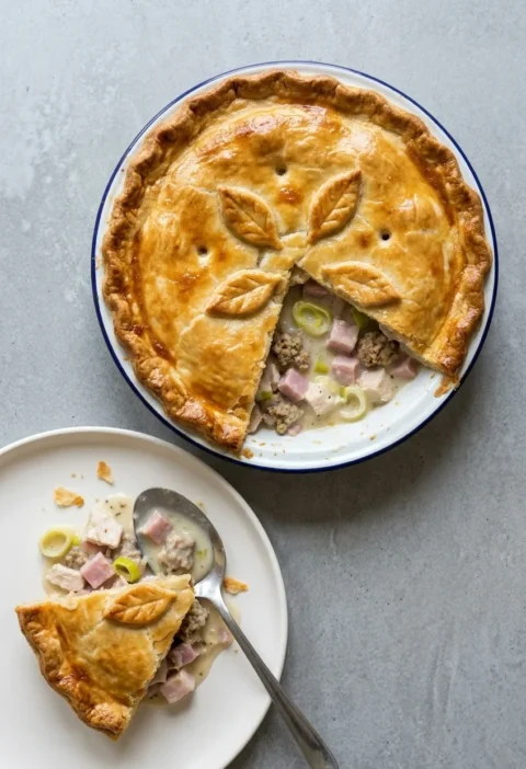 Golden turkey and ham pie with pastry leaves in an enamel dish, with a slice on a plate showing turkey, ham, leeks and stuffing in a creamy sauce