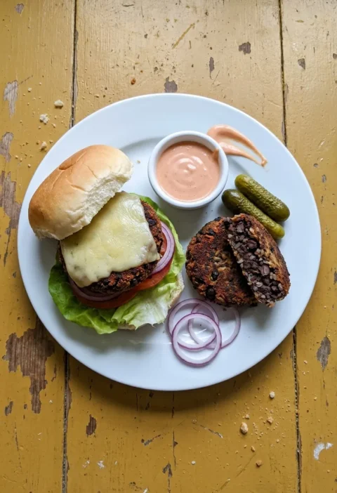 Black bean veggie burger with melted cheese, a halved bean patty, gherkins and a ramekin of burger sauce on a white plate, on a worn yellow table