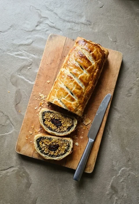 Golden veggie Wellington on a wooden board with two slices cut, showing mushroom and spinach around a chestnut and lentil filling with a prune